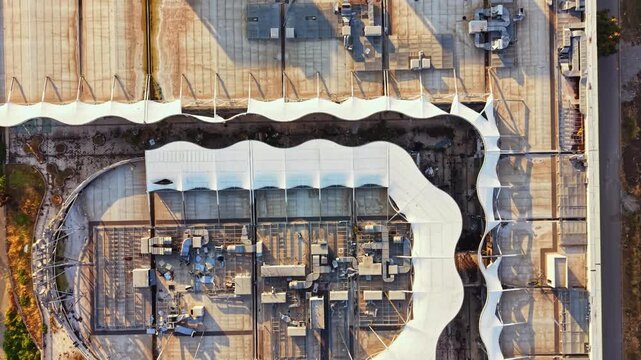 A large building with a curved roof is seen from above. The roof features several sections and is surrounded by other structures in the urban landscape during the day.