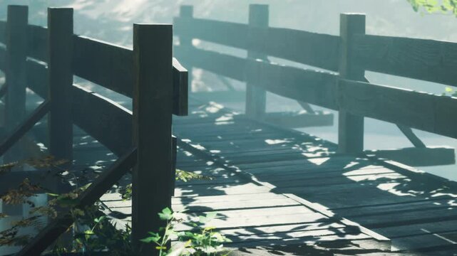 A wooden bridge crosses a small path among trees. Mist hangs low in the air, and shadows fall across the bridge. Sunlight filters through the branches.