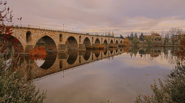 Masonry Pointed arch Stone bridge (Puente de Piedra) across the river Duero in Zamora, Castile-Leon, Spain. Reflection in the water. Pedestrians walking on bridge.