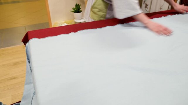 Hand adjusts white fabric with red border. Material appears soft and thick. Background shows wooden floor and curtains. Scene captures gentle handling of textile. Lighting is warm and even