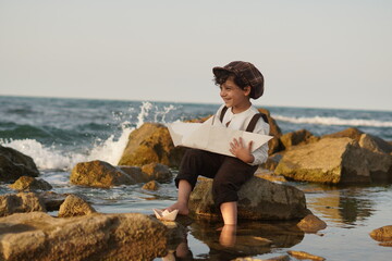 Happy little boy in vintage outfit playing with a big paper boat by the sea © Samane
