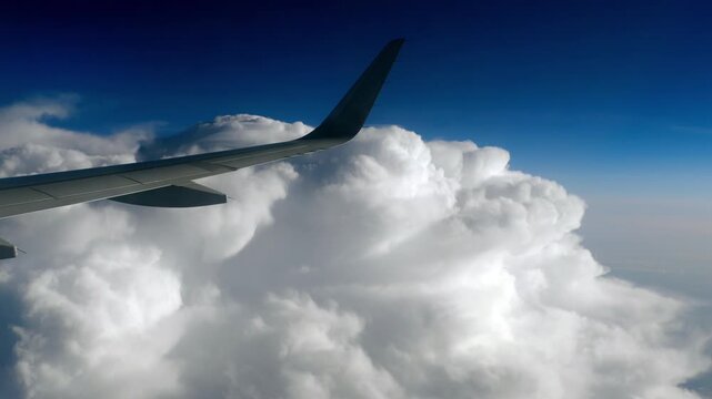 Airplane wing flying high above massive white cumulus clouds in bright blue sky during daytime, passenger window view.
