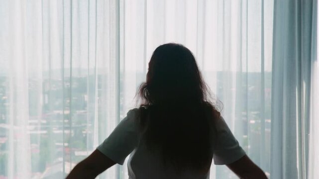 Back lit silhouette of woman with arms raised triumphantly gazing through floor to ceiling windows at expansive city skyline from high rise apartment or hotel room.