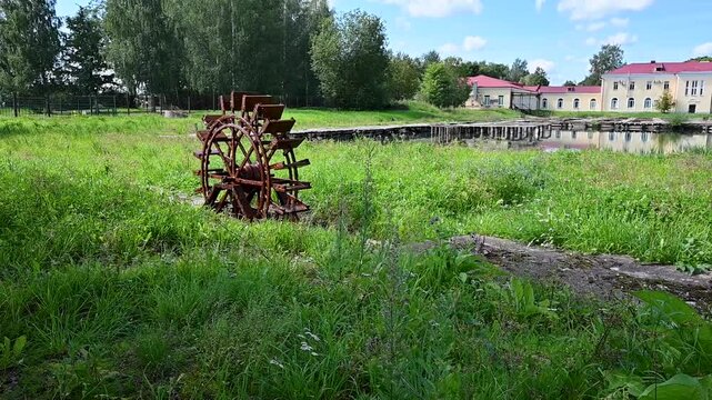 Rusted waterwheel stands in green grassy field. Wooden structure nearby overlooks calm, shallow pond. Lush trees frame background under bright sky. Red-roofed building visible beyond the water