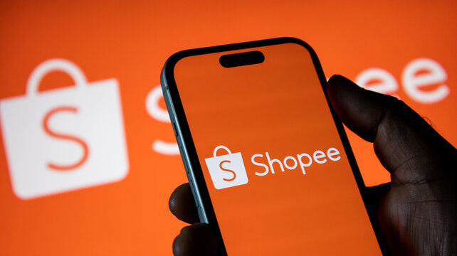 Hand holding smartphone with Shopee logo against orange background and shopping bag icon.