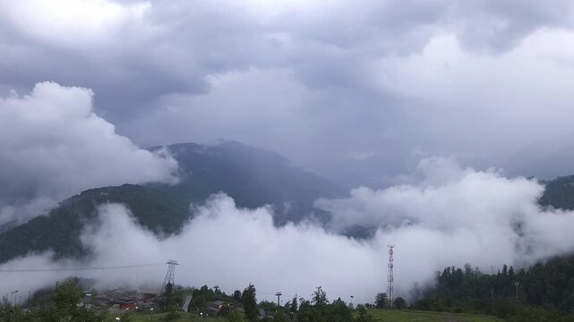 Majestic panoramic view of a mountain landscape with thick fog and low clouds slowly clearing to reveal a dense green forest and distant snowy peaks under a dramatic, overcast sky