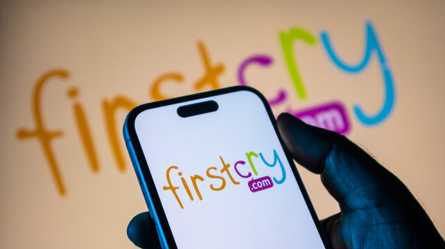 A close-up view of firstcry com website logo on a smartphone screen, with a larger logo blurred in the background.