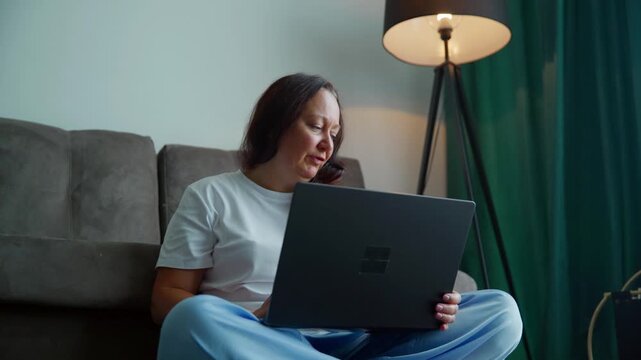 Woman relaxing on gray sofa, working on laptop in contemporary apartment with ambient floor lamp, green curtains, natural lighting from window.