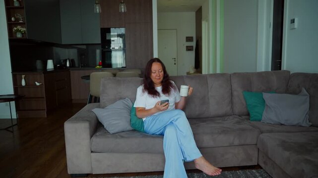 Woman sitting on gray sectional sofa holding coffee cup, scrolling smartphone in contemporary open plan apartment with kitchen visible.