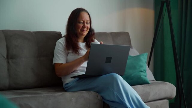 Woman sitting on gray couch with turquoise pillow, working on laptop computer in casual home setting with green decor elements.