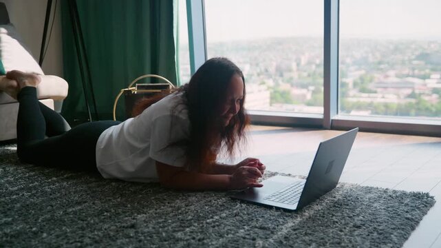 Woman reclines on carpet floor with laptop in modern high rise apartment overlooking sprawling cityscape through floor to ceiling windows, relaxed work posture.