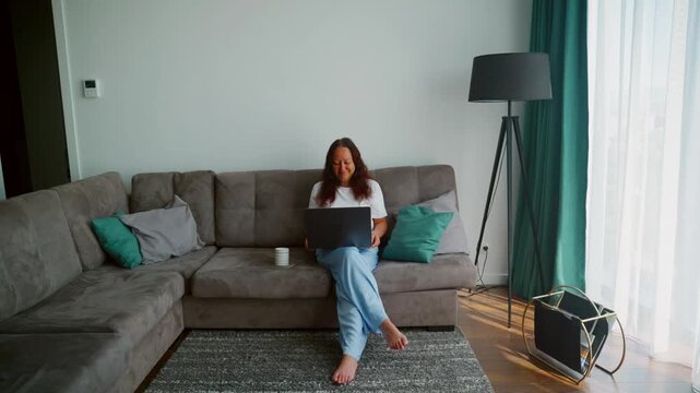 Woman seated on gray sectional couch with laptop in contemporary living room with teal accents, floor lamp, and natural light from windows.