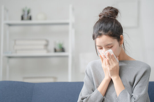 Seasonal illness concept. Young woman blowing her nose with a tissue while sitting at home, representing cold, flu and allergy symptoms. Cold, flu, allergy, tissue, health.