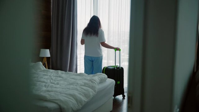 Female traveler holding black suitcase stands near bright window in modern hotel bedroom, preparing for journey. Scene captures travel readiness, tourism, and accommodation lifestyle.