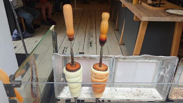 Static shot of rotating chimney cakes on vertical spits baking and caramelizing at street food stall window with wooden handles and metal tray daytime market scene. Trdelnik, a typical Prague pastry