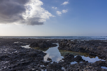 Fototapeta premium Dramatic dark storm clouds contrasting with bright sky over volcanic coast pools