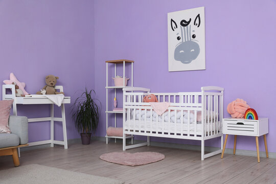 Baby crib, shelving unit, changing table and bedside table near violet wall in stylish childrens room