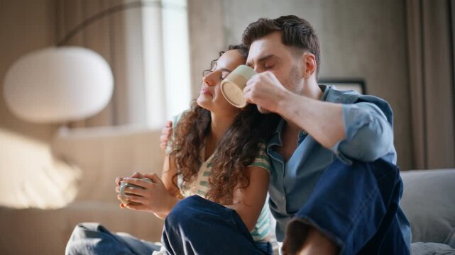 Interracial couple cuddling coffee cups home. Man woman weekend morning sofa.