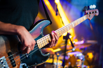 Obraz premium Close-up of a bass guitarist playing during a live concert with colorful stage lighting and blurred drum kit in the background.