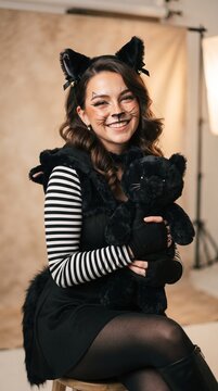 Halloween A young woman dressed as a playful black cat, complete with ears and whiskers, smiles warmly while holding a stuffed teddy bear in a studio setting, embodying a spirit of fun and whimsy.