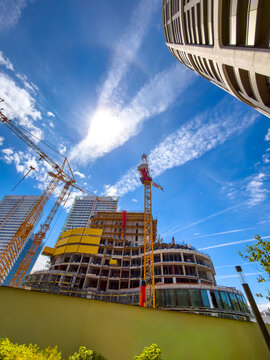Construction site under bright sky. Workers build a new structure surrounded by cranes and tall buildings in a city. Clouds fill the blue sky above.