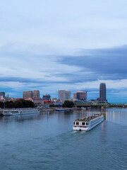 Obraz premium Cityscape with boat on river at dusk. A boat moves along the river as buildings rise above the water in a city at dusk. The sky is filled with clouds.