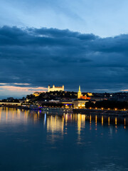 Fototapeta premium Skyline of the city with lights at dusk. A view of the city skyline shows buildings and a castle lit up in the evening. The water reflects the lights.