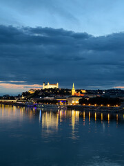Naklejka premium City skyline view at night with river. Lights reflect on the river as buildings shine on the hill. The scene captures a city at night with clouds above.