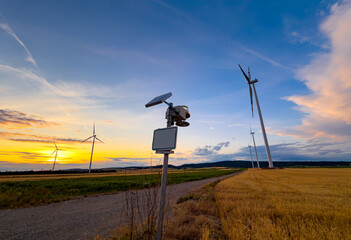 Wind turbines stand in a field at sunset. Wind turbines stand in a field at sunset. A weather station with sensors is nearby. © Анна Терелюк