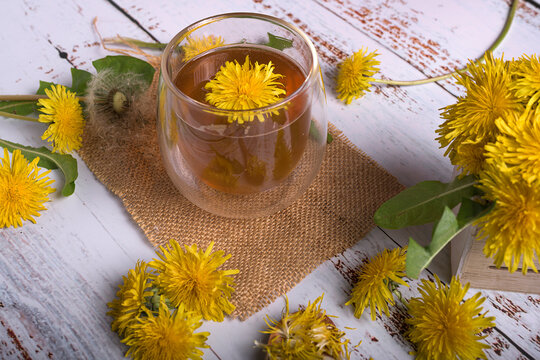 Top view of a glass mug filled with healthy dandelion tea decorated with a yellow flower and mint leaves on a rustic burlap cloth background