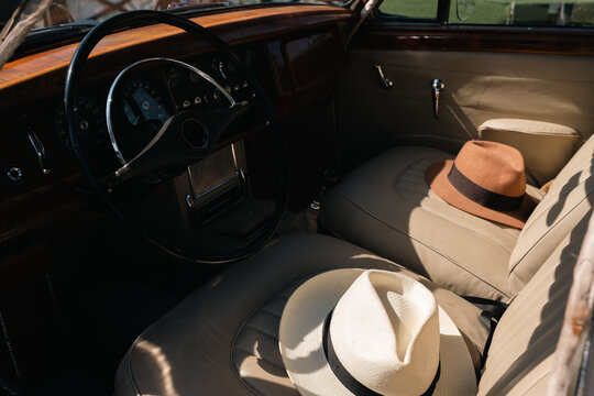 Interior of a vintage classic car with leather seats, a wooden dashboard, and two stylish straw hats resting on the passenger side.