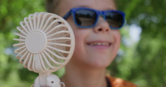Happy boy in sunglasses against green trees in park looking away and blowing wind from portable fan in hand.