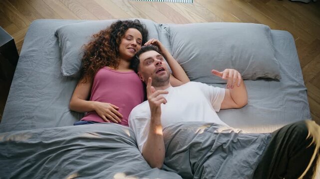 Happy couple laughing bed morning. Woman resting head boyfriend chest chatting.