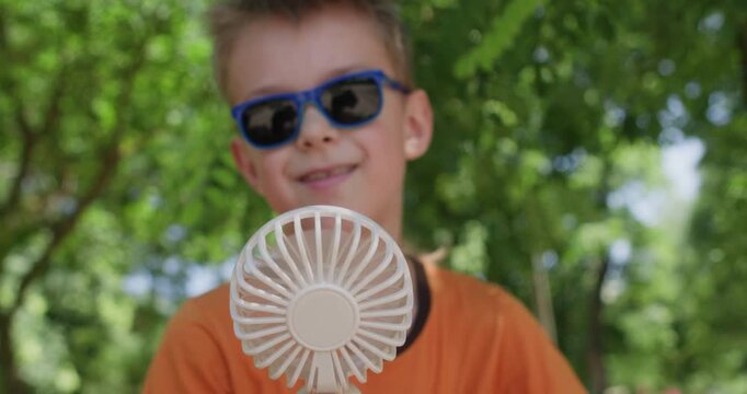 A boy wearing sunglasses lets a portable fan blow on his face in the summer heat, rubbing his forehead and hair with his hand.