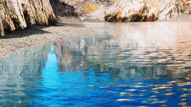Water shows clear reflections of rocks and the sky at a beach surrounded by cliffs. Sand and pebbles line the shore where waves gently touch the land.