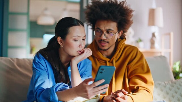 Couple reading shocking news phone home sofa. Closeup worried multiethnic pair.