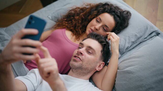 Couple relaxing bed taking selfie phone. Woman resting on boyfriend cozy morning
