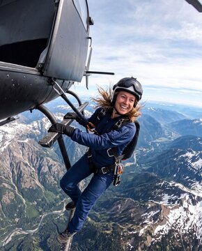 Smiling female daredevil hangs from a helicopter skid above mountains before a skydive jump. Concept of adventure, courage, and overcoming fear.