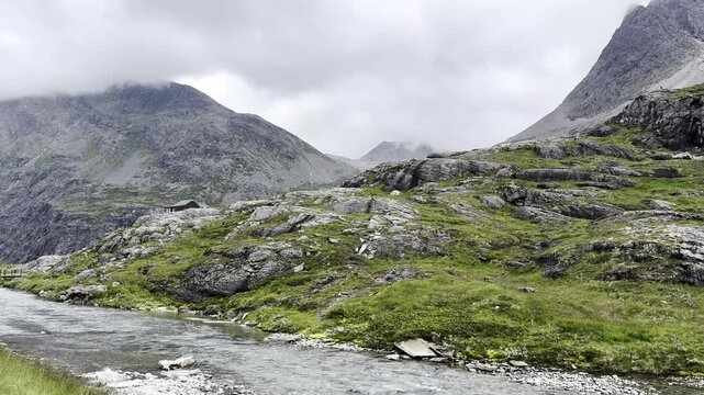 Spectacular view of the famous Trollstigen mountain pass in Norway with a winding river flowing through the valley below. Dramatic mountains, low clouds, mist drifting across the peaks, and lush Scand
