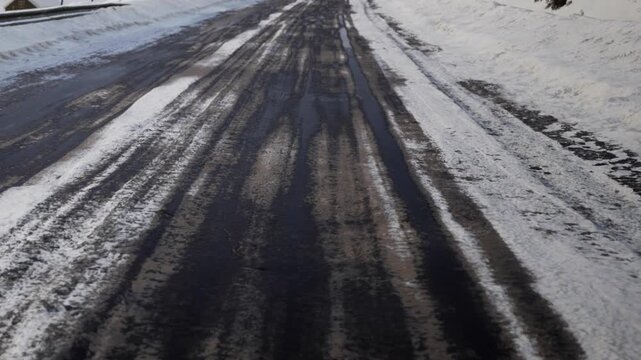 A road runs through a rural area covered in snow. Tire tracks are visible on the wet surface. The scene shows a cold winter day with snow piled along the edges of the road.