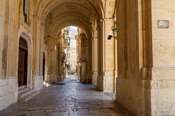 Obraz premium A shaded stone archway leads through a cobblestone passage lined with ornate limestone architecture in Valletta, Malta