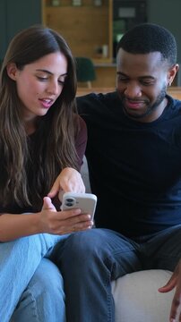 A happy multi-ethnic couple relaxes on a sofa, sharing a smartphone and enjoying content together. They are smiling, interacting, and showcasing connection and shared digital experience at home.