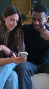 A happy diverse couple relaxing on a couch, sharing and looking at content on a mobile phone. They smile and interact, showing connection and modern digital lifestyle at home.