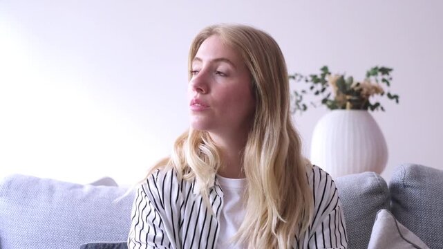 A thoughtful young blonde woman sits comfortably on a modern sofa at home, her gaze fixed off-camera. Her pensive expression conveys deep thought, contemplation, or perhaps waiting, capturing a moment
