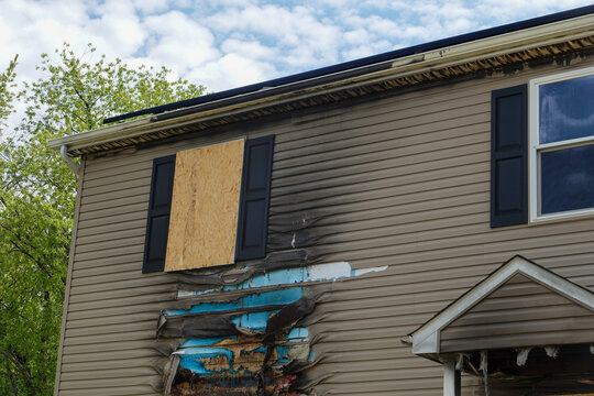 Boarded up window on a house destroyed by fire with charred and melted vinyl siding, eves and soffit