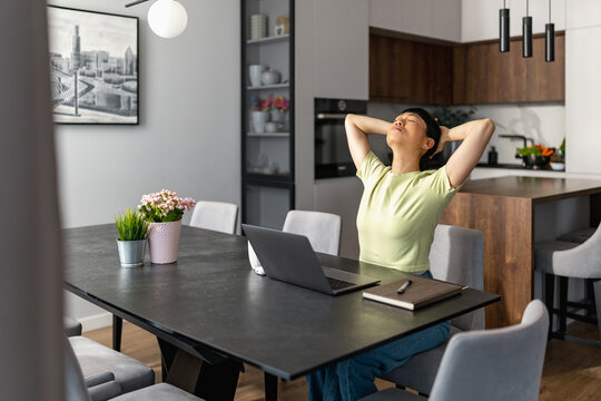 Japanese woman stretching at kitchen table while working from home with laptop and notebook
