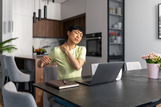 Japanese woman rubbing sore neck while working from home at laptop in modern kitchen