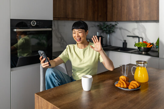 Japanese woman smiling and waving during a video call in a modern kitchen with breakfast