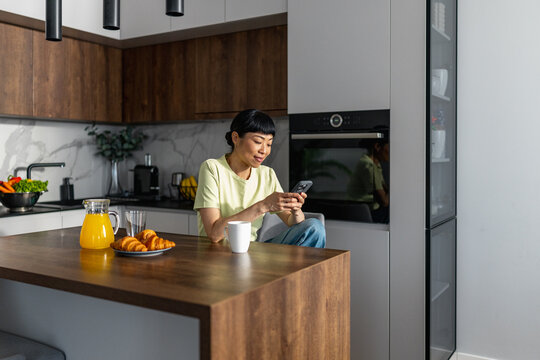 Japanese woman checking phone while sitting on kitchen island with breakfast and coffee