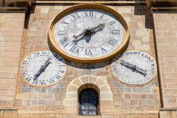 Fototapeta premium An ornate church clock face with Roman numerals, flanked by smaller date and day dials, adorns a stone tower in Valletta, Malta